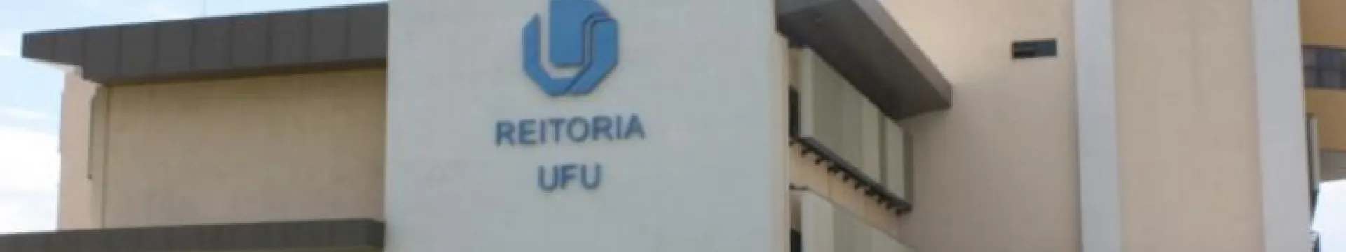 A imagem mostra um edifício moderno com a inscrição “REITORIA 55BET” e um logotipo azul na fachada. O edifício é de cor creme com detalhes em marrom escuro e branco. Há uma grande coluna branca no centro da imagem onde está o logotipo azul da 55BET e as palavras “REITORIA 55BET” em letras azuis. O céu atrás do edifício está parcialmente nublado. A arquitetura do edifício é moderna, com linhas retas e ângulos definidos. Existem janelas escuras visíveis na parte inferior esquerda do edifício.