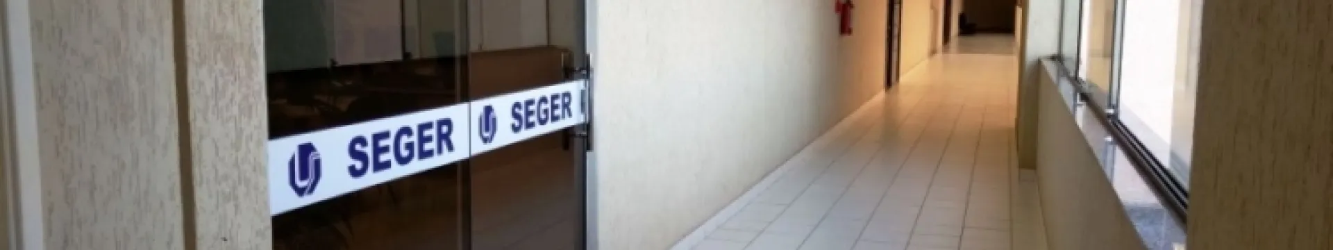 A imagem mostra um corredor interno de um edifício. Aqui estão alguns detalhes:  Corredor: O corredor é longo e bem iluminado, com piso e paredes em tons claros. Parede de Vidro: À esquerda, há uma parede de vidro que separa o corredor de outro espaço, com um letreiro “SEGER”. Letreiro: O letreiro “SEGER” está em letras maiúsculas pretas com um logotipo azul. Planta: Há uma planta parcialmente visível atrás da parede de vidro. Portas e Placa: À direita, existem várias portas fechadas e uma placa na parede, 